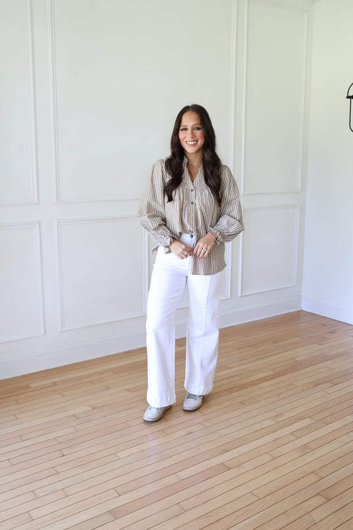 A woman stands in a bright room with white paneled walls and light wood floors, wearing a beige and white vertically striped button-down shirt, white wide-leg pants, and white sneakers.