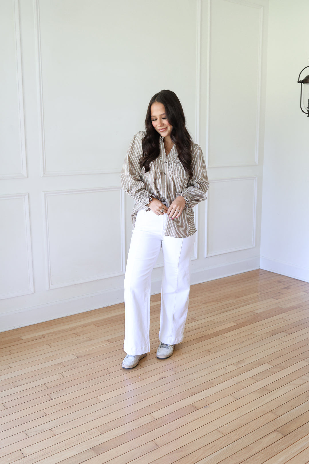 A woman stands in a bright room with white paneled walls and light wood floors, wearing a beige and white vertically striped button-down shirt, white wide-leg pants, and white sneakers.