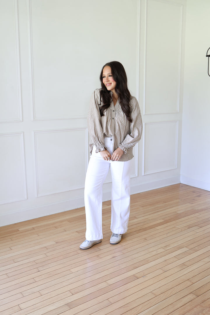 A woman stands in a bright room with white paneled walls and light wood floors, wearing a beige and white vertically striped button-up shirt, white wide-leg pants, and silver shoes.