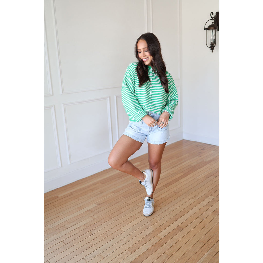 Woman in a green striped sweater and white shorts posing in a room with wooden flooring and white walls.