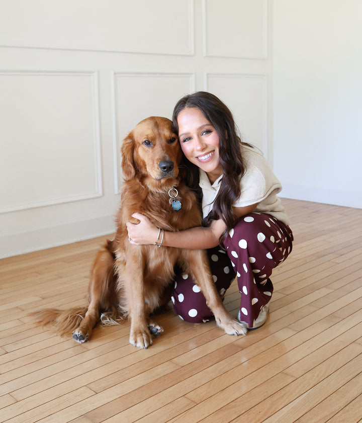 A smiling woman crouches on a light wood floor and hugs a golden retriever dog. She is wearing a light beige short-sleeve top and burgundy pants with white polka dots.