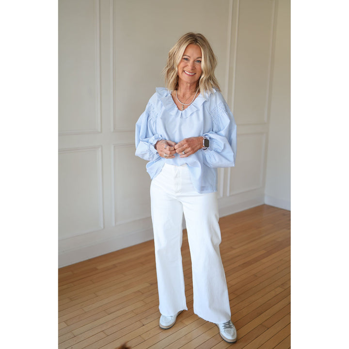 Woman wearing a light blue blouse and white pants standing in a room with wooden flooring and white paneled walls.