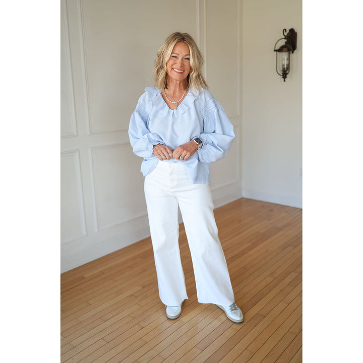 Woman wearing a light blue blouse and white pants standing in a room with wooden flooring and a wall sconce.