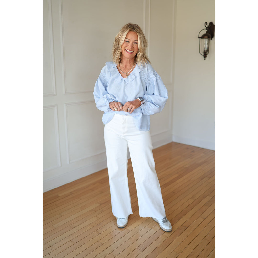 Woman wearing a light blue blouse and white pants standing in a room with wooden flooring and white walls.