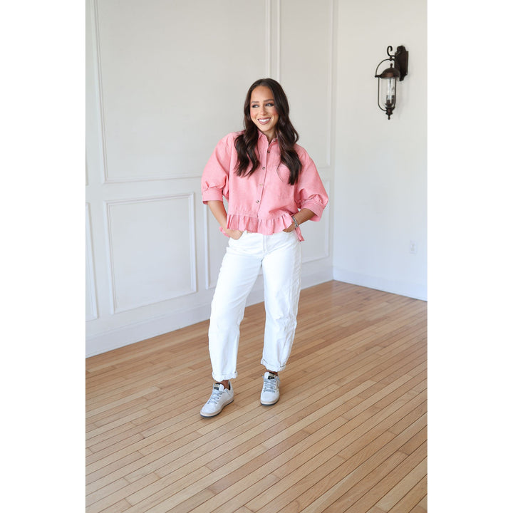 Woman wearing a pink blouse and white pants standing in a room with wooden flooring and white walls.