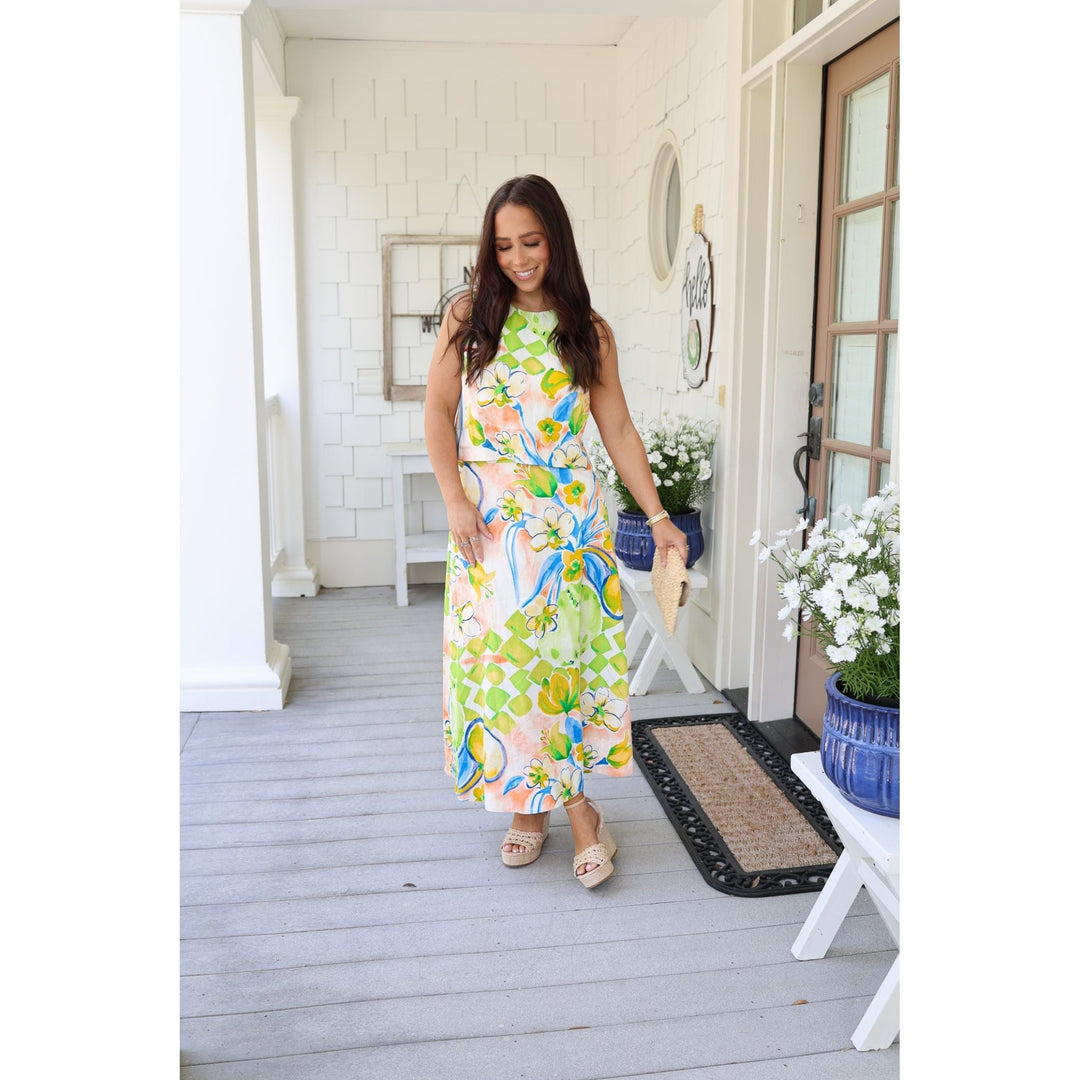 Woman in a colorful dress standing on a porch with flowers and a chair.