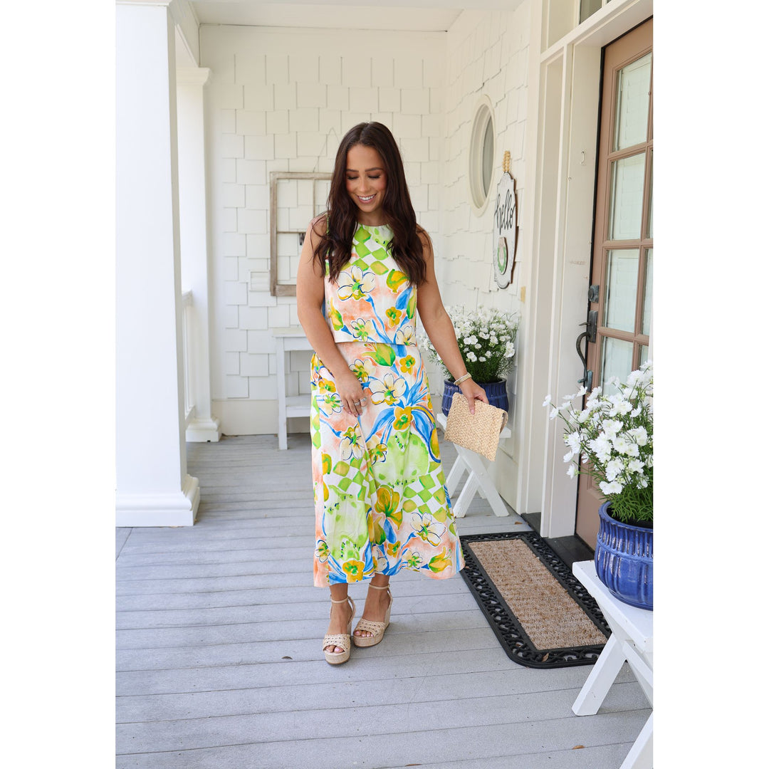 Woman in a colorful floral dress standing on a porch with flowers and a plant in the background.