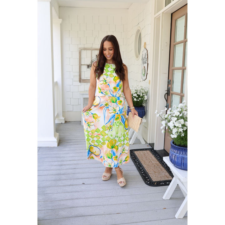 Woman in a colorful dress standing on a porch with flowers and a chair in the background.