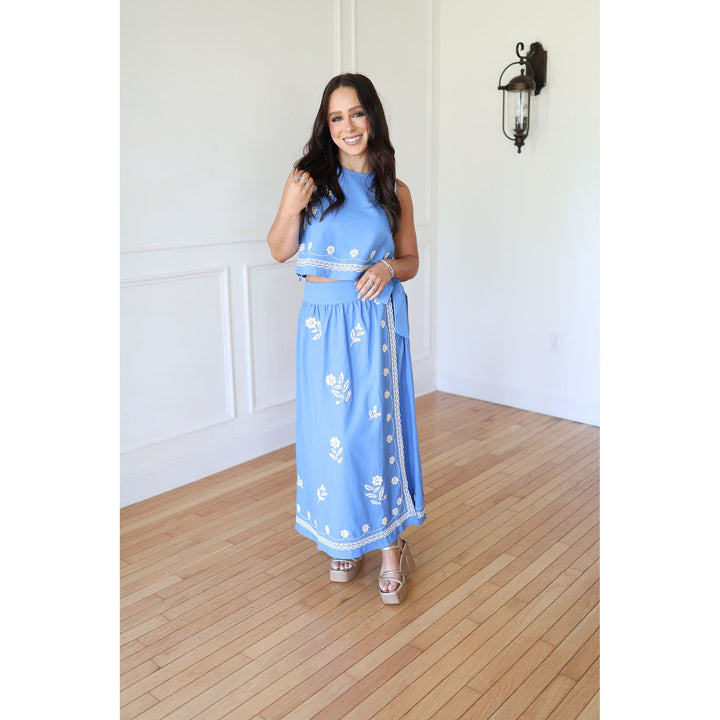 Woman wearing a blue outfit with white patterns in a room with wooden flooring and white walls.