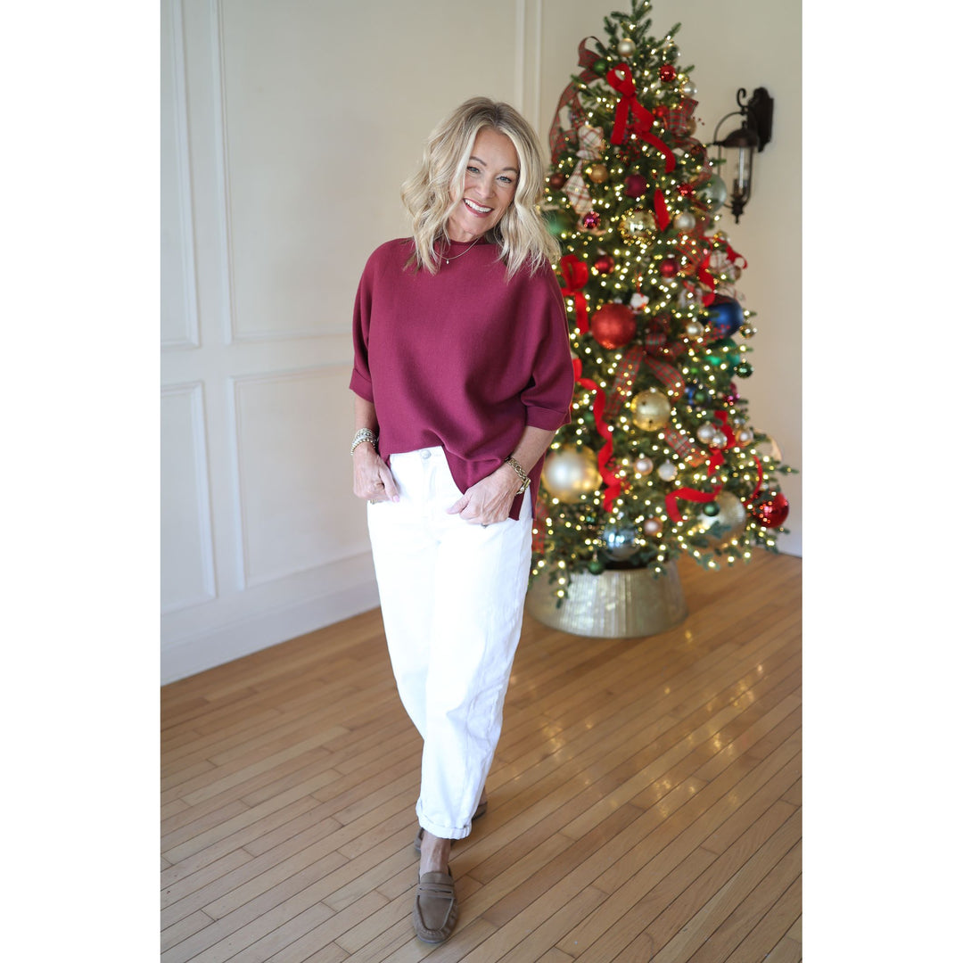 Woman in a maroon sweater and white pants standing in front of a decorated Christmas tree.