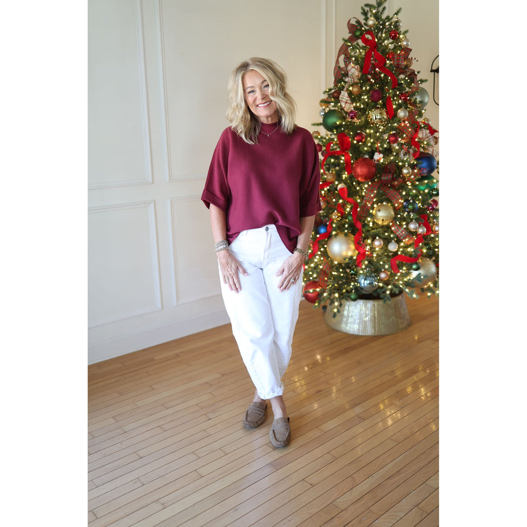 Woman in a burgundy sweater and white pants standing in front of a decorated Christmas tree.