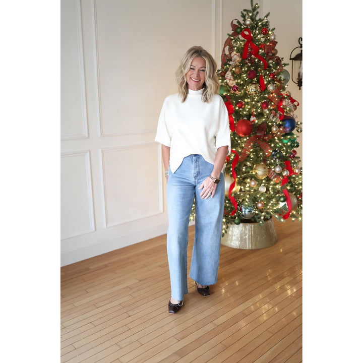 Woman in a white top and blue jeans standing next to a decorated Christmas tree indoors.