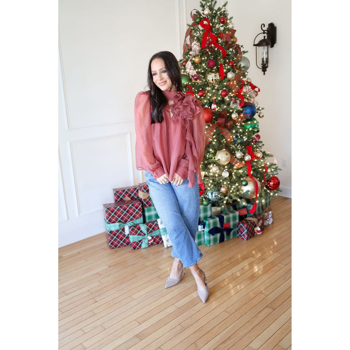 Woman in a pink blouse and blue jeans standing in front of a decorated Christmas tree with presents underneath.