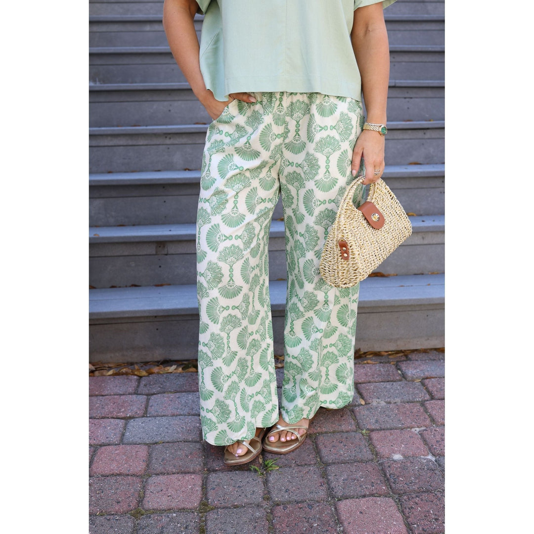 Person wearing green patterned pants and holding a straw bag against a metal shutter background