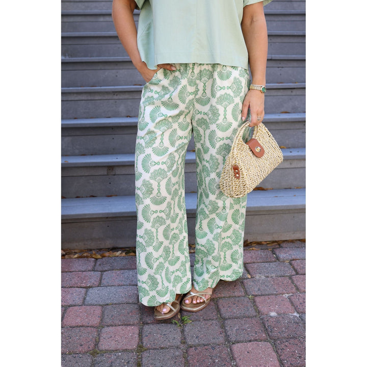 Person wearing green patterned pants and holding a straw bag against a metal shutter background