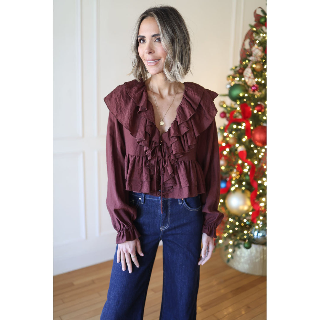 Woman wearing a burgundy ruffled blouse and blue jeans standing in front of a decorated Christmas tree.