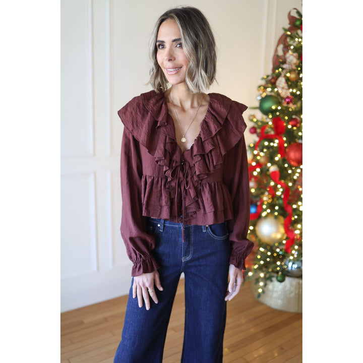 Woman wearing a burgundy ruffled blouse and blue jeans standing in front of a decorated Christmas tree.