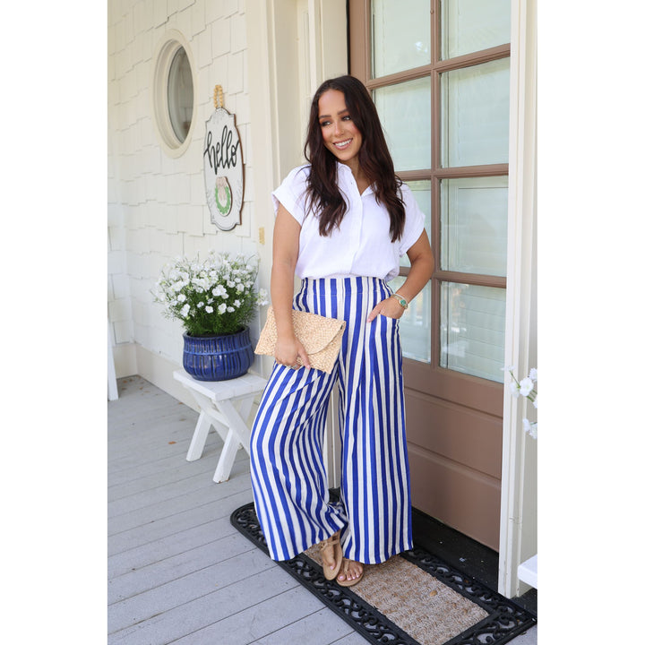 Woman in white shirt and blue striped pants standing on a porch with a 'Hello' sign and flowers in the background.