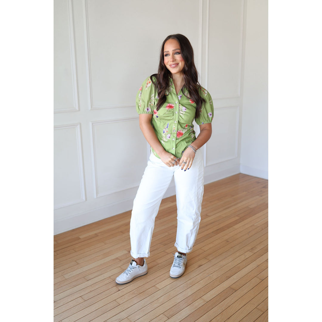 Woman wearing a green floral shirt and white pants standing on a wooden floor.