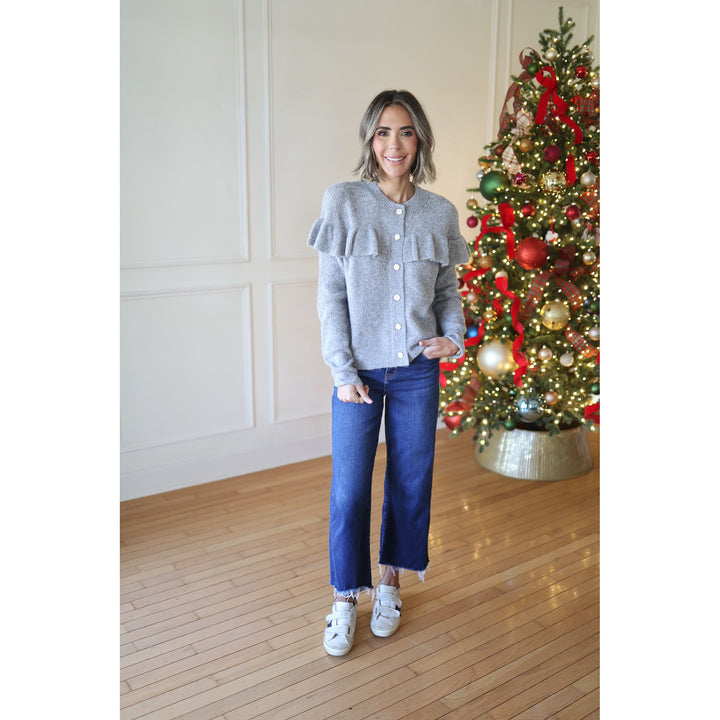 Woman standing in front of a decorated Christmas tree wearing a gray sweater and blue jeans.
