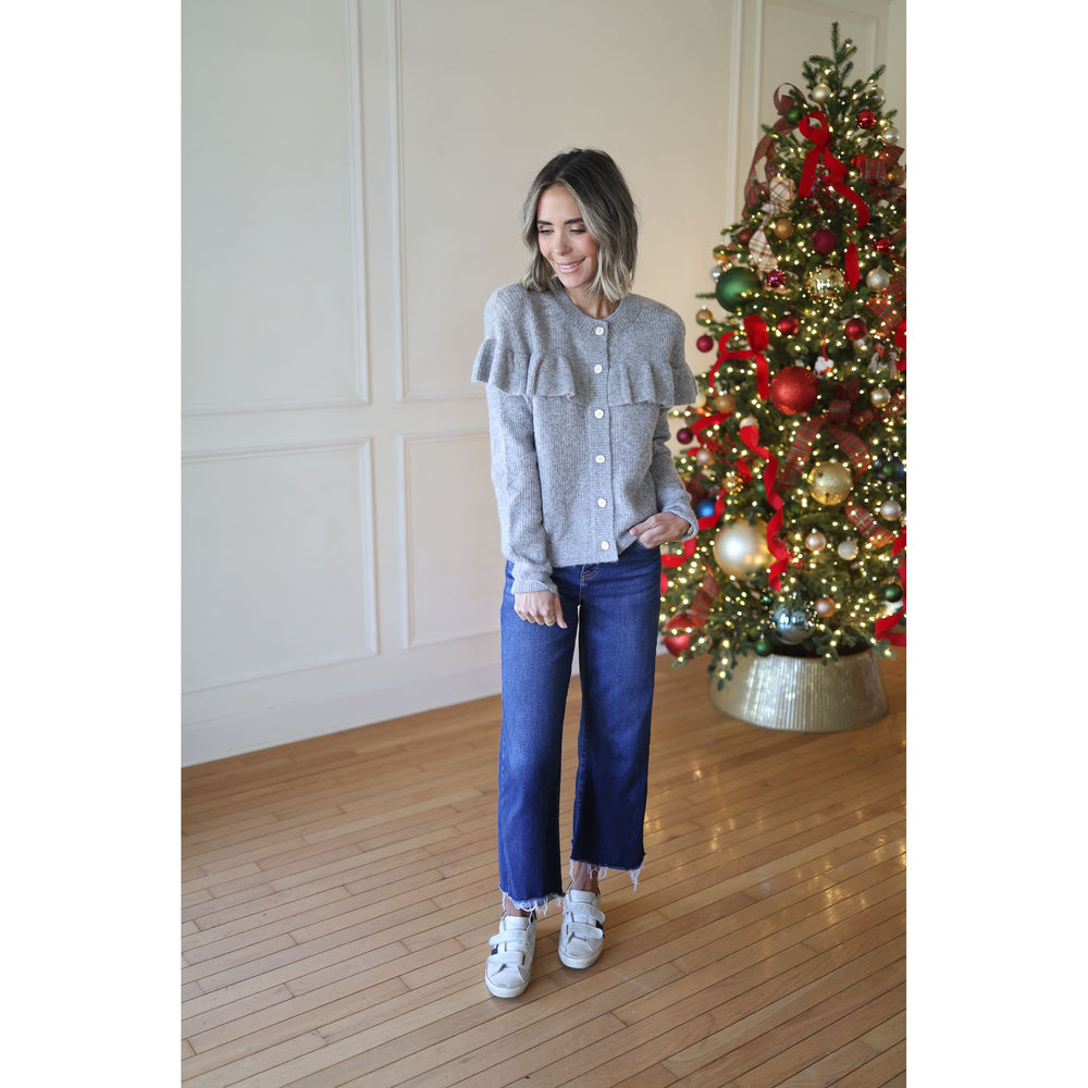 Woman standing in front of a decorated Christmas tree wearing a gray sweater and blue jeans.