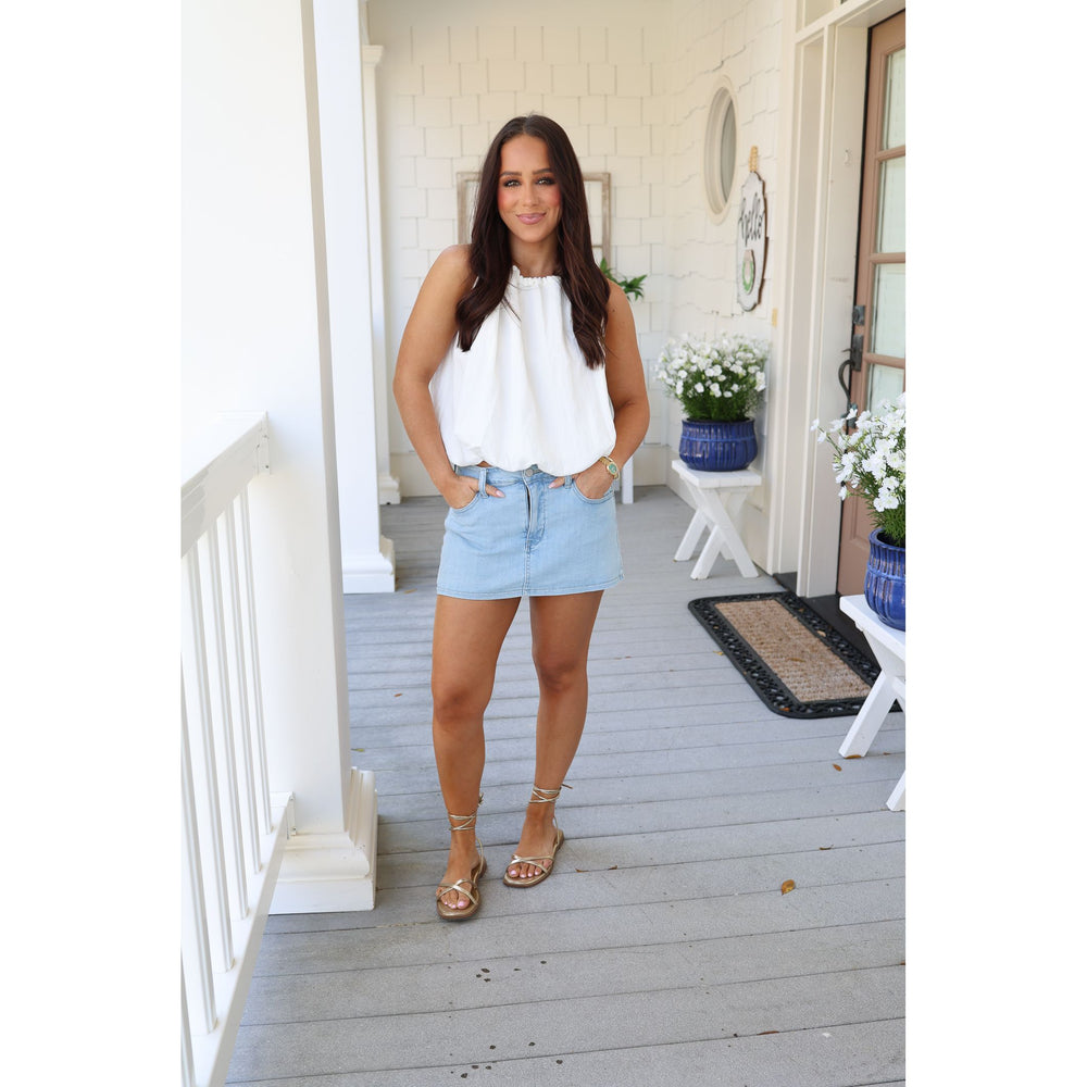 Woman standing on a porch wearing a white top and light blue shorts.