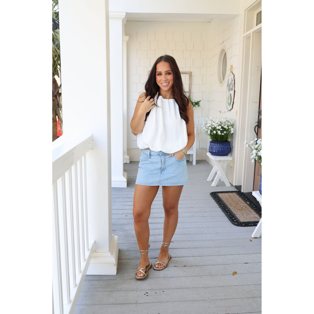 Woman standing on a porch wearing a white blouse and light blue shorts.