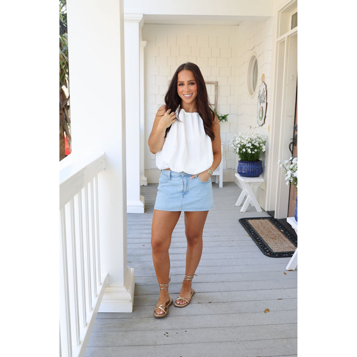 Woman standing on a porch wearing a white blouse and light blue shorts.