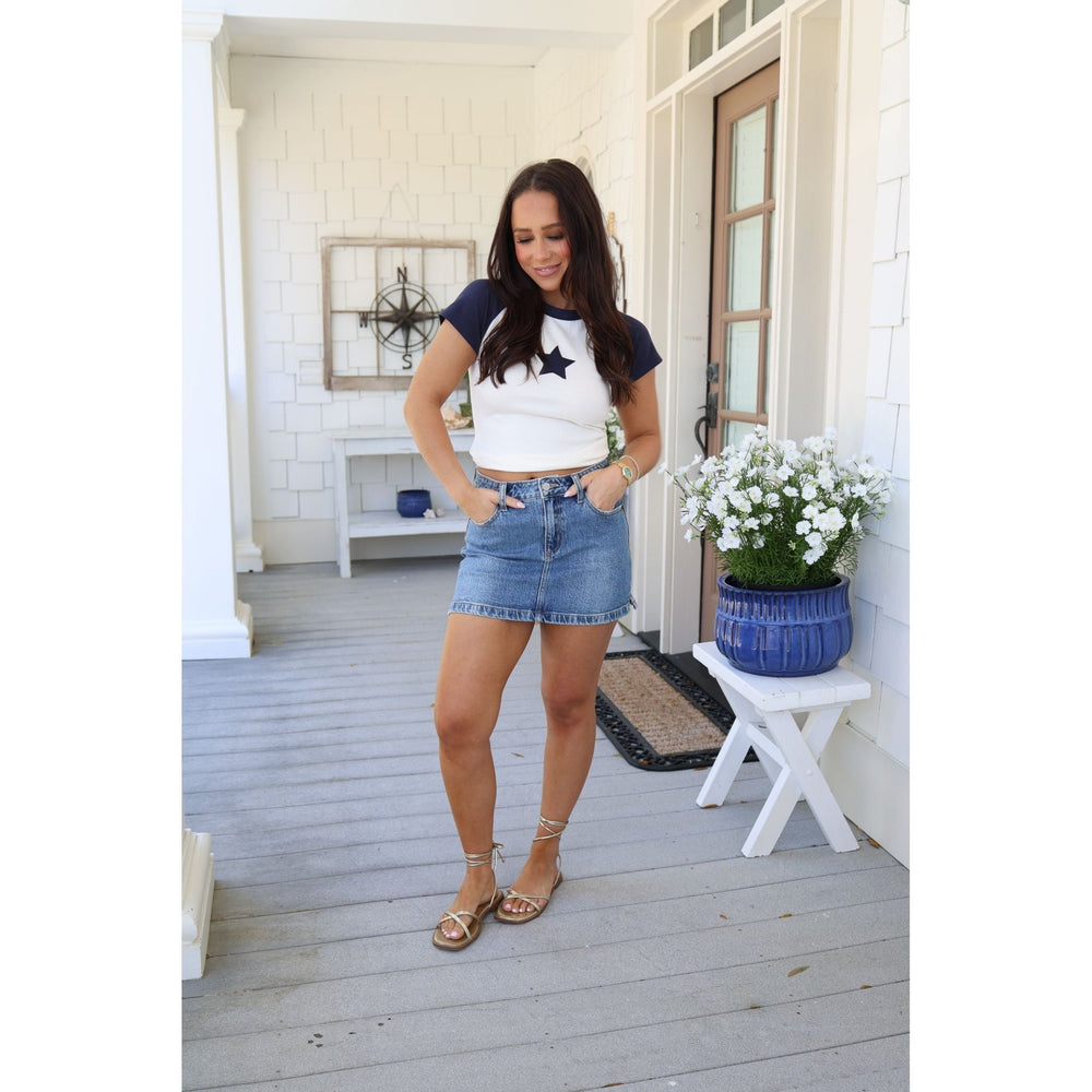 Woman standing on a porch wearing a white crop top with a blue star and denim shorts.