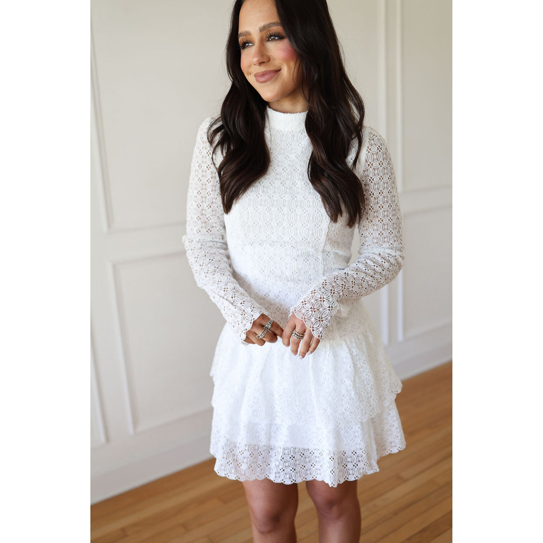 Woman wearing a white lace dress standing against a neutral wall.