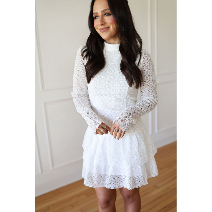 Woman wearing a white lace dress standing against a neutral wall.
