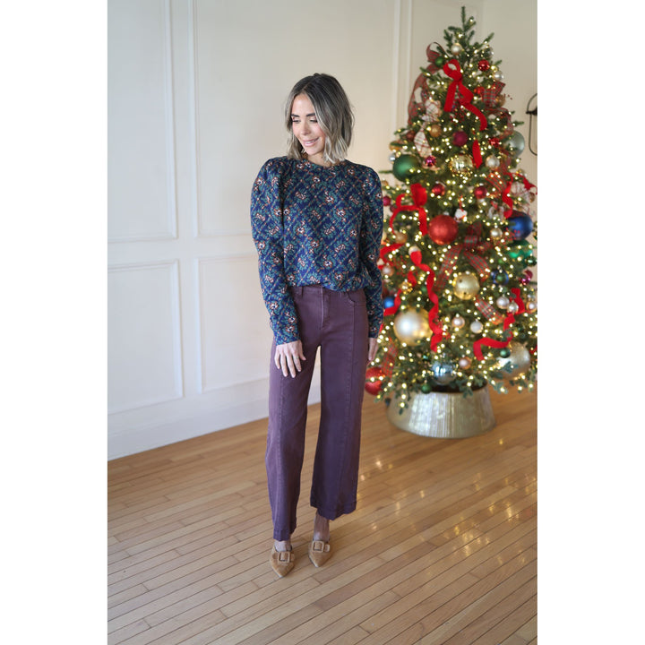 Woman in a blue patterned sweater and purple pants standing in front of a decorated Christmas tree.