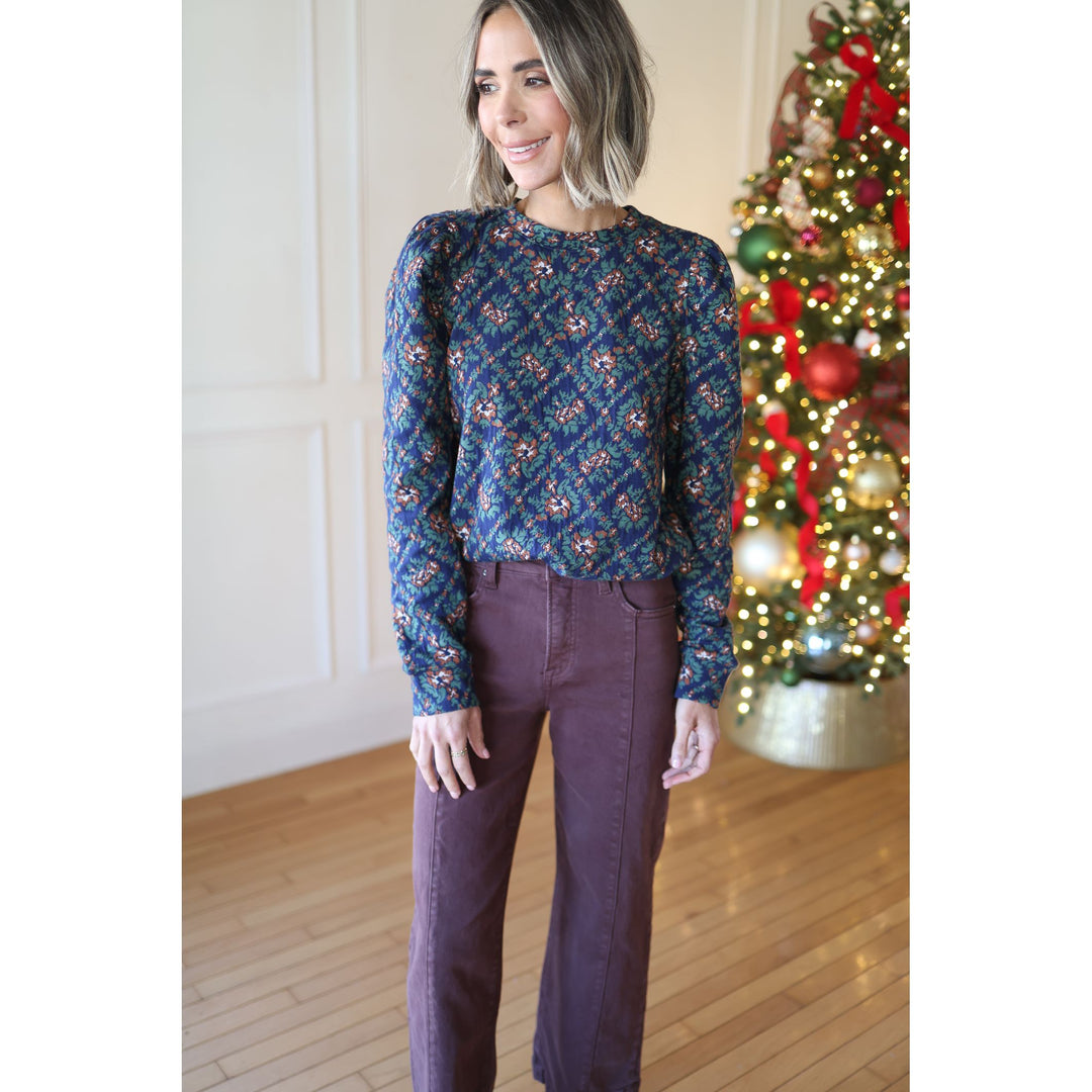 Woman wearing a patterned blue top and purple pants standing in front of a decorated Christmas tree.