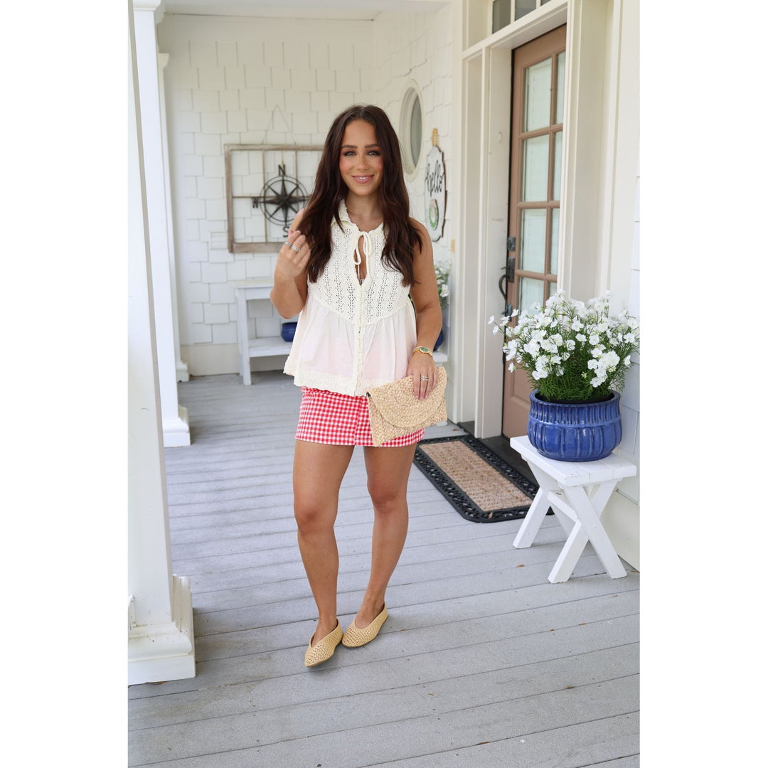 Woman standing on a porch wearing a white top and red checkered skirt, holding a straw bag.