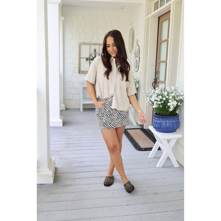 Woman standing on a porch wearing a beige top and zebra print skirt.