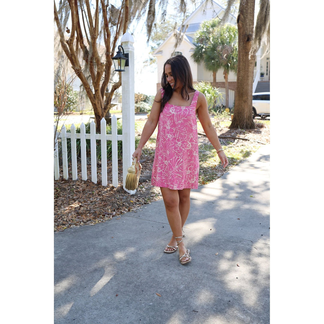 Woman in a pink dress standing on a sidewalk with a white picket fence and house in the background.