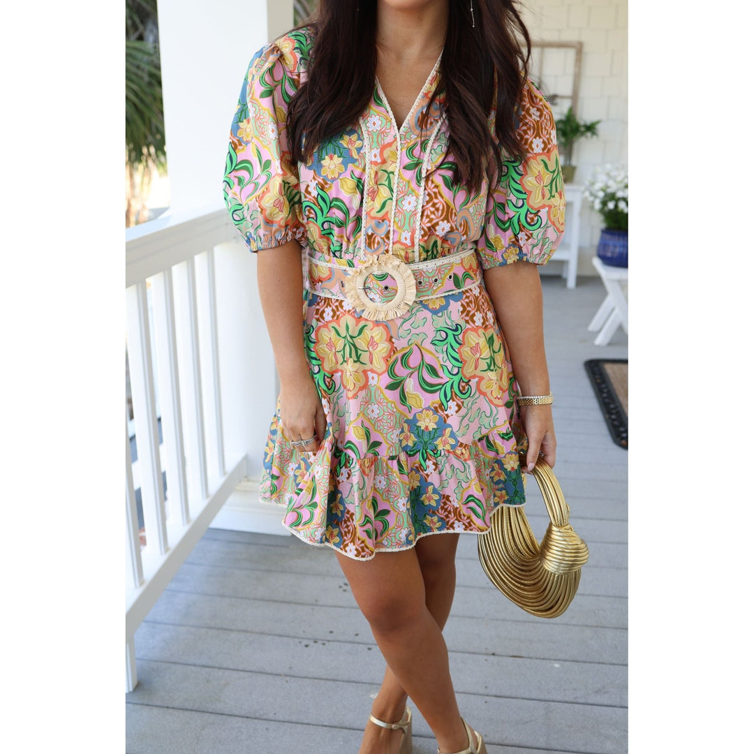 Woman wearing a colorful floral dress with a belt and gold purse on a porch.