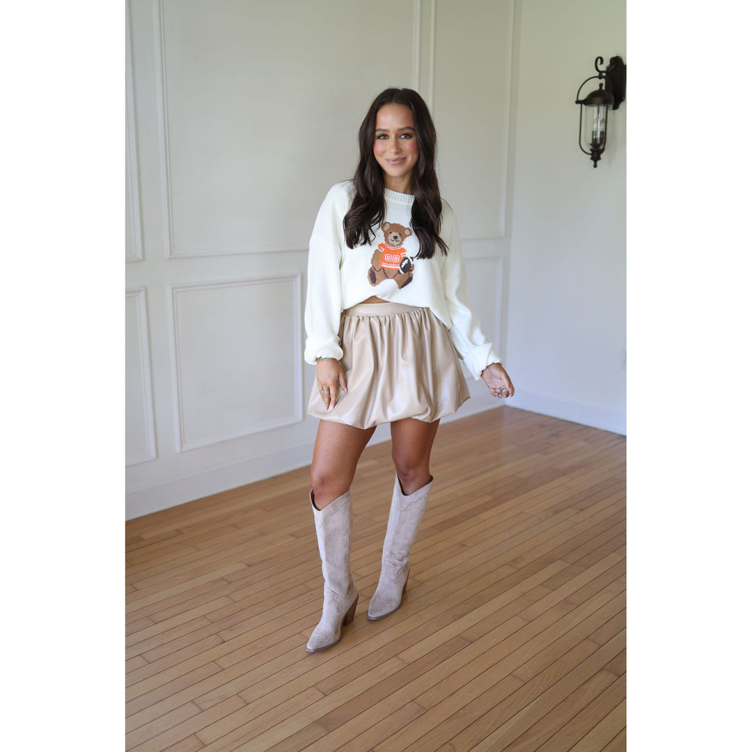 Woman wearing a white sweater with a teddy bear design, beige skirt, and white knee-high boots