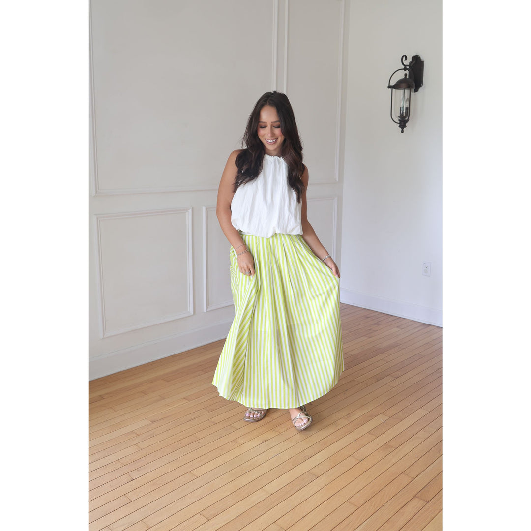 Woman wearing a white top and yellow striped skirt standing in a room with wooden flooring and white walls.