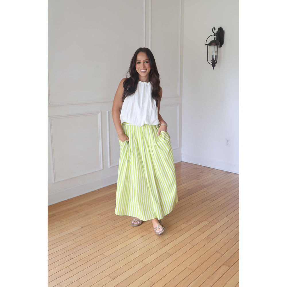 Woman wearing a white top and green striped skirt standing in a room with wooden flooring and white walls.
