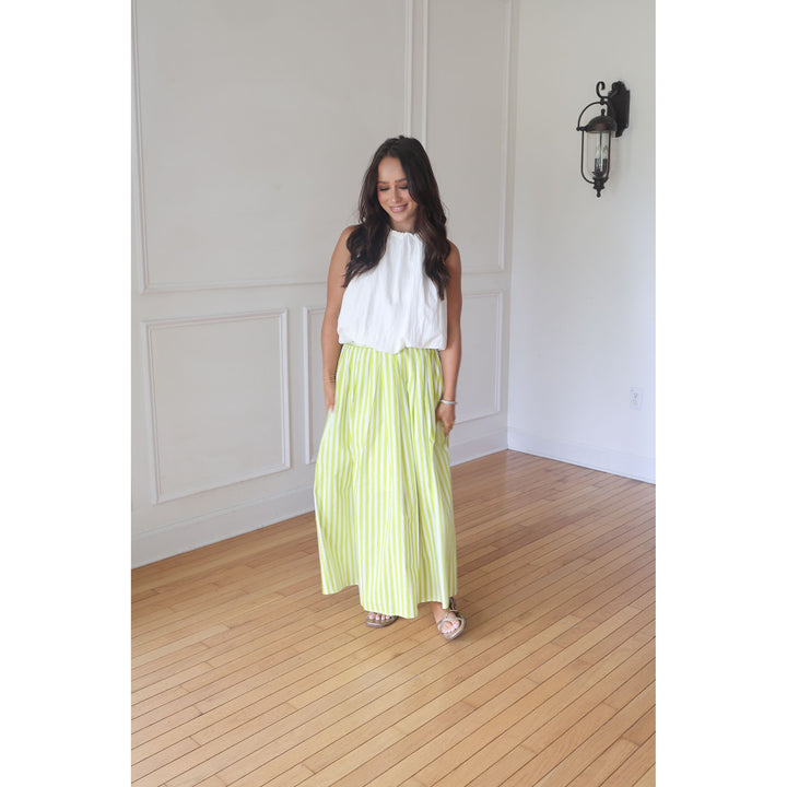 Woman wearing a white top and green striped skirt standing in a room with wooden flooring and white walls.