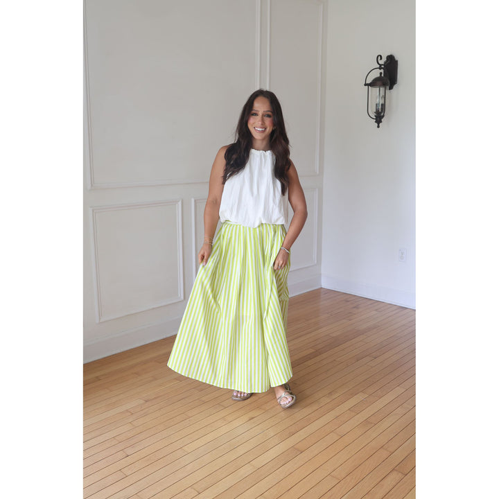Woman wearing a white top and green striped skirt standing in a room with wooden flooring and white walls.