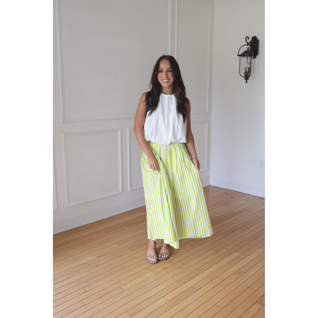Woman wearing a white top and green striped skirt standing in a room with wooden flooring and white walls.