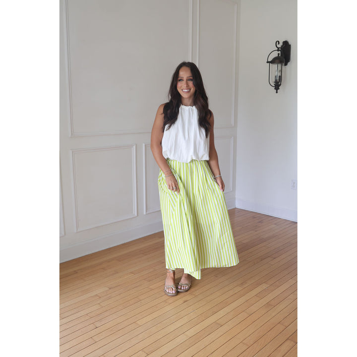 Woman wearing a white top and green striped skirt standing in a room with wooden flooring and white walls.