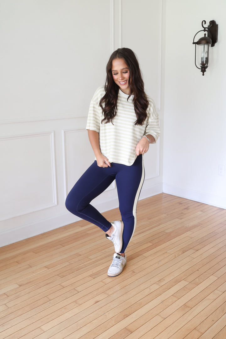 A woman posing wearing a cream and navy striped long-sleeve top, navy blue leggings with a white side stripe, and white sneakers.