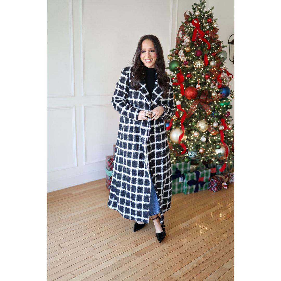 Woman in a checkered coat standing in front of a decorated Christmas tree with presents underneath.