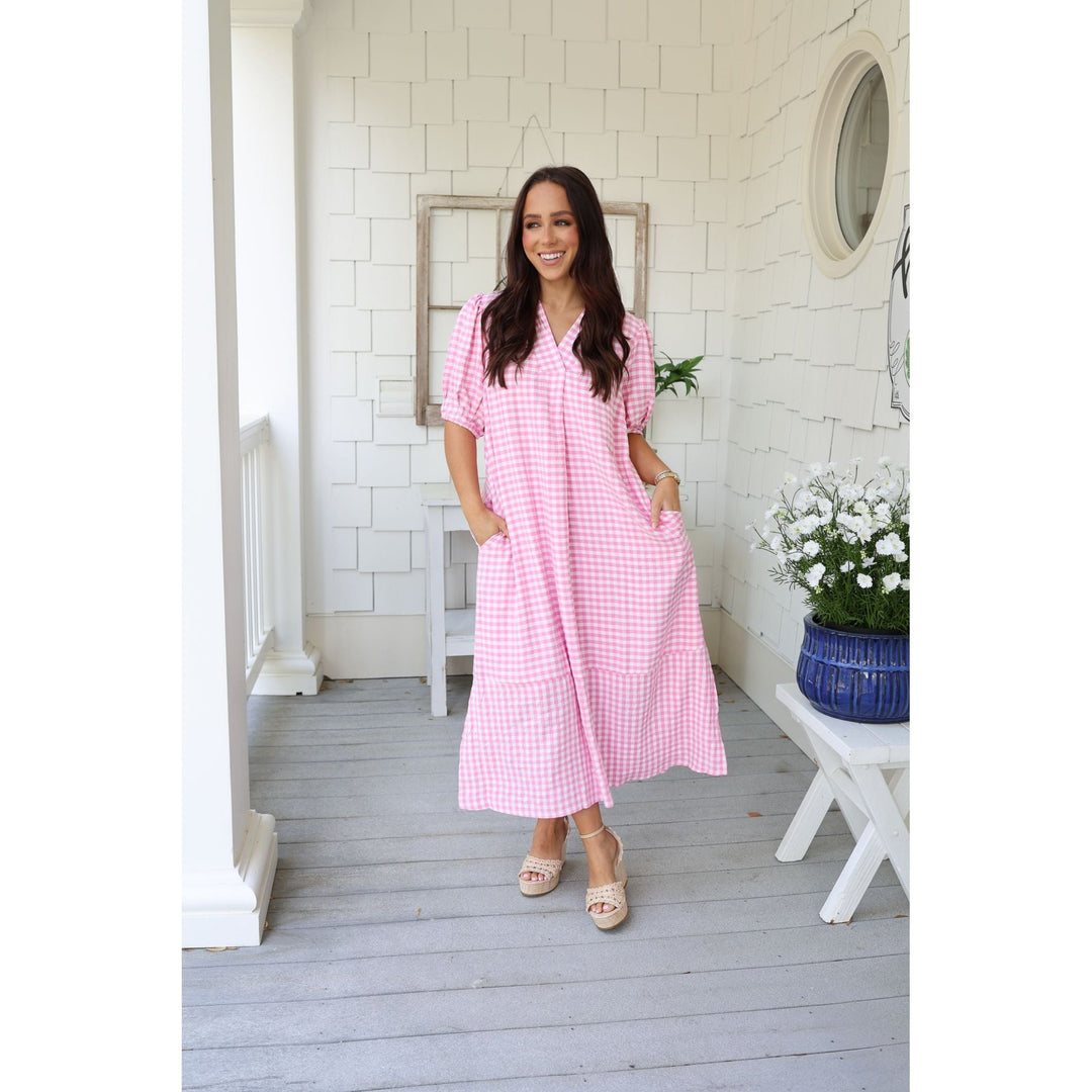 Woman in a pink checkered dress standing on a porch with white walls and a small table with flowers.