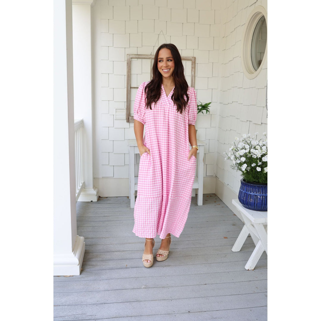Woman in a pink checkered dress standing on a porch with white walls and a plant in the background.