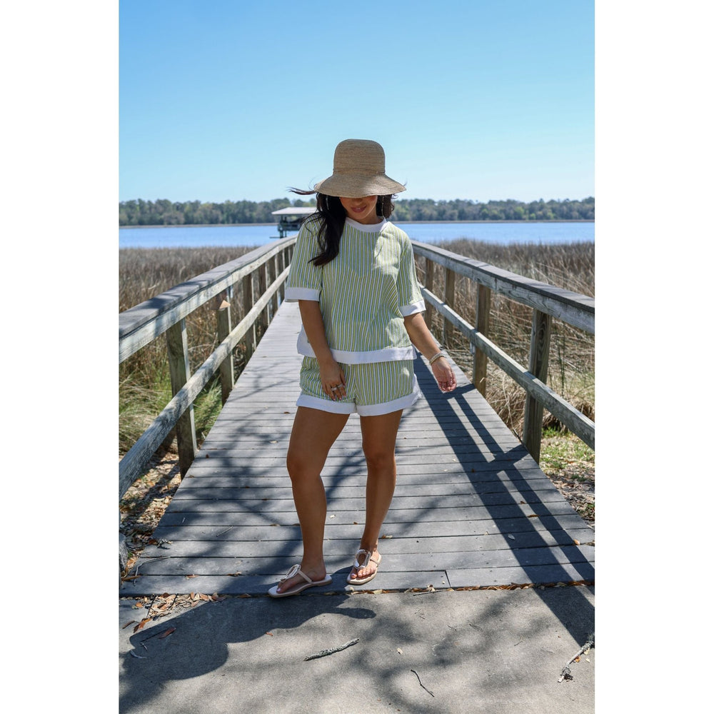 Woman standing on a wooden boardwalk by a body of water, wearing a green and white striped shirt and shorts.