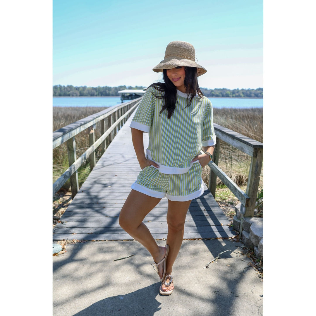 Woman in a striped shirt and shorts standing on a wooden boardwalk by a body of water.